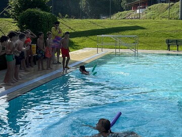 Zwei Schülerinnen bei Übungen mit Schwimmnudel im Pool, Mitschüler stehen am Beckenrand